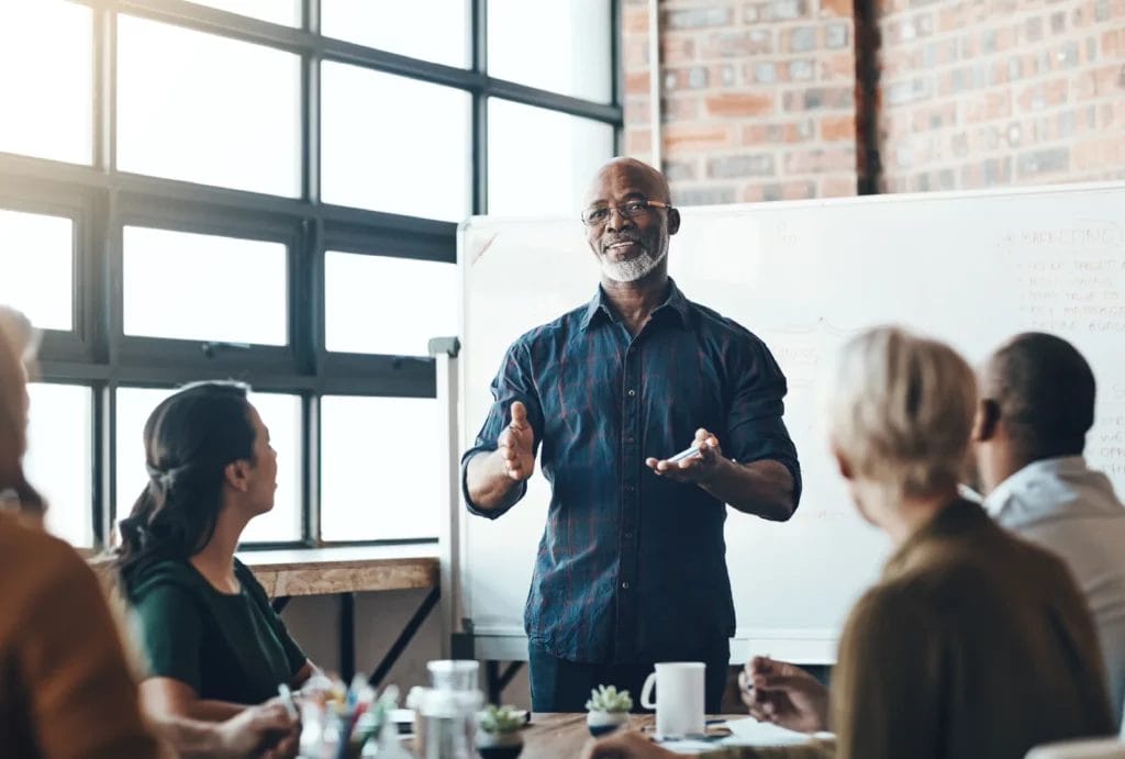 A business coach presents in front of a group of employees at a corporation, using a whiteboard to outline key concepts.