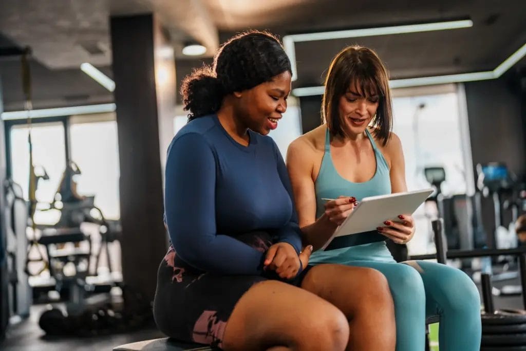 Smiling personal trainer presenting a training plan on a digital tablet to a cheerful woman sitting in a gym, fostering motivation and support
