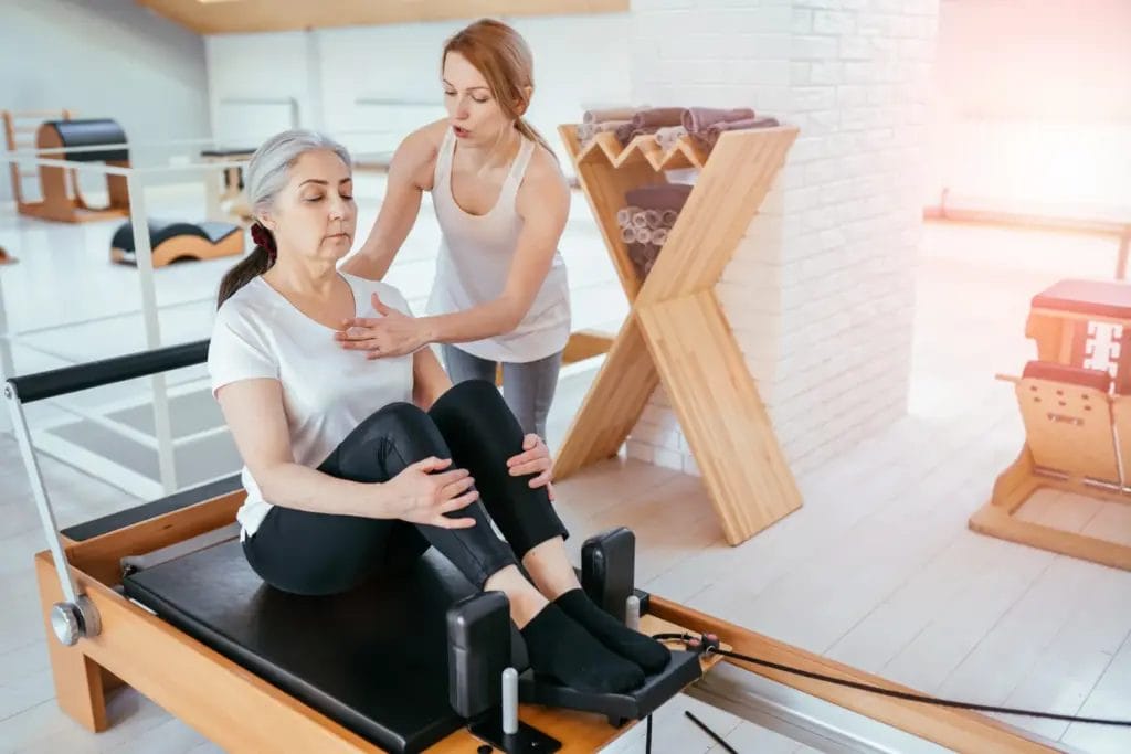 A Pilates instructor guides a client's breathwork and form while the client sits on a Pilates reformer in a Pilates studio.
