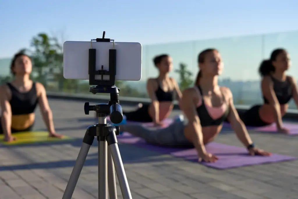 A smartphone set up on a tripod filming an outdoor yoga class of students practicing upward dog on yoga mats on a rooftop.