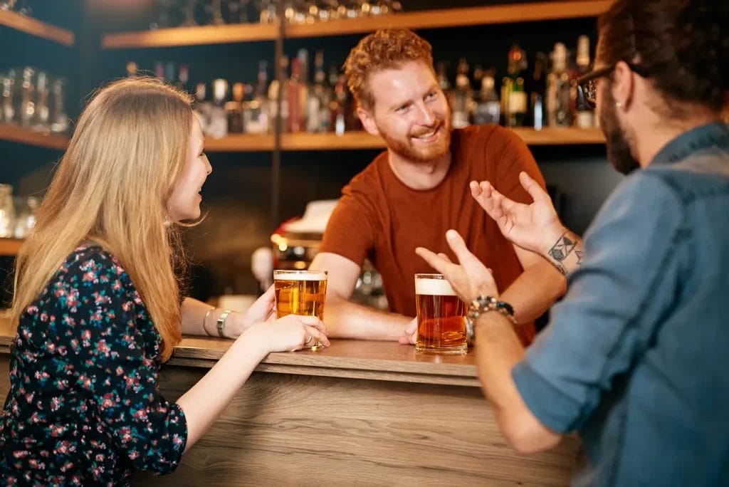 A bartender smiles at two patrons while serving them glasses of beer.