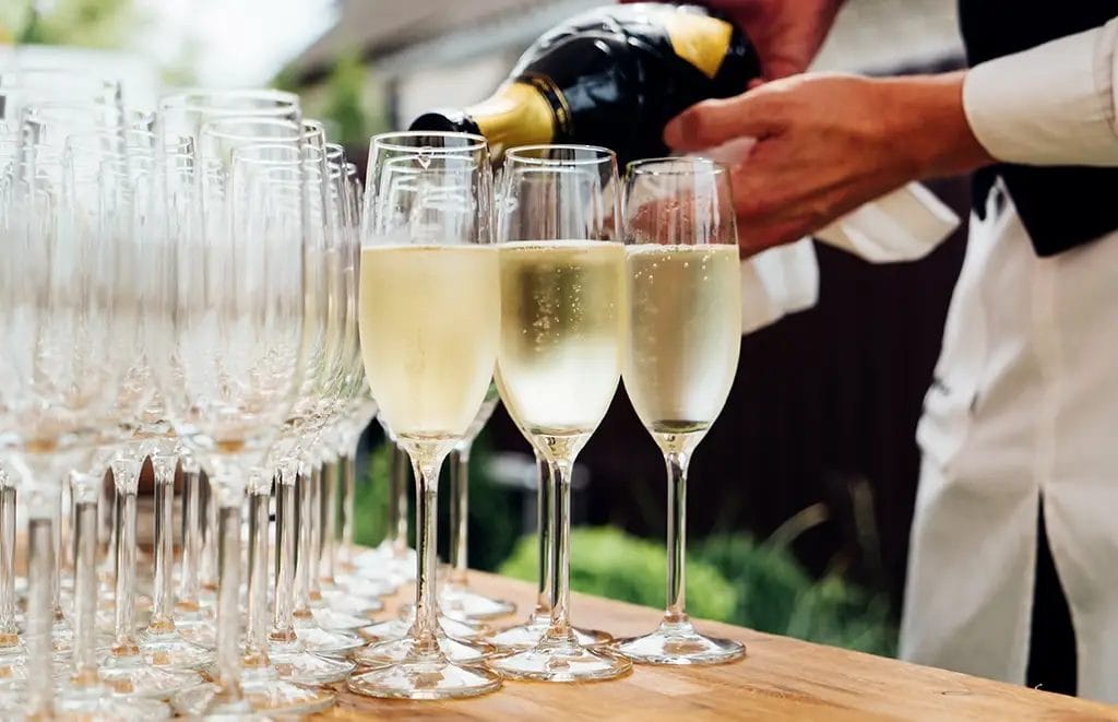 A bartender pouring a bottle of champagne into flutes outdoors.