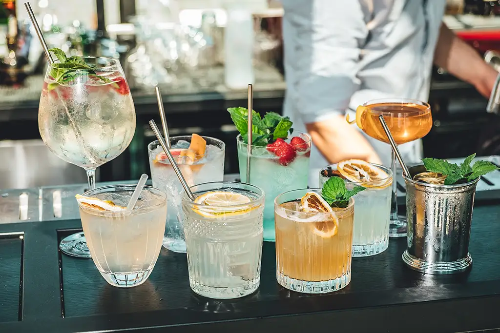 Nine different cocktails garnished with herbs and citruses sitting on an outdoor bar.