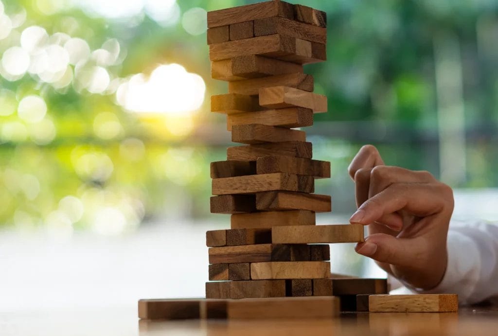 Hand pulling blocks out of a wobbly block tower, symbolizing risk.
