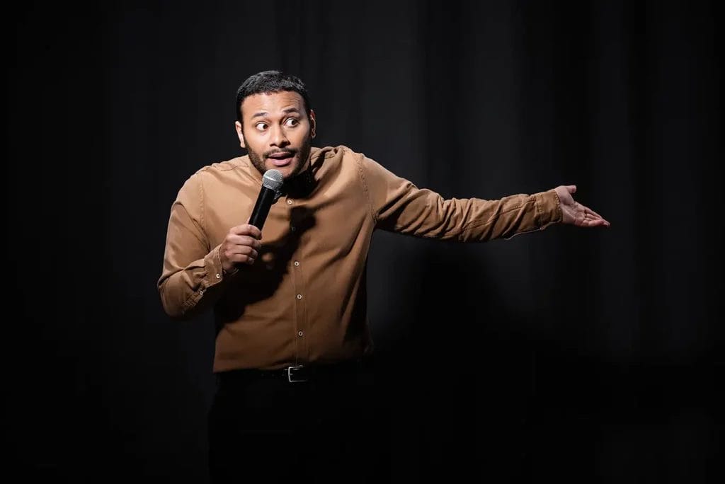 A man in a brown button-up shirt performs on stage in front of a black backdrop.