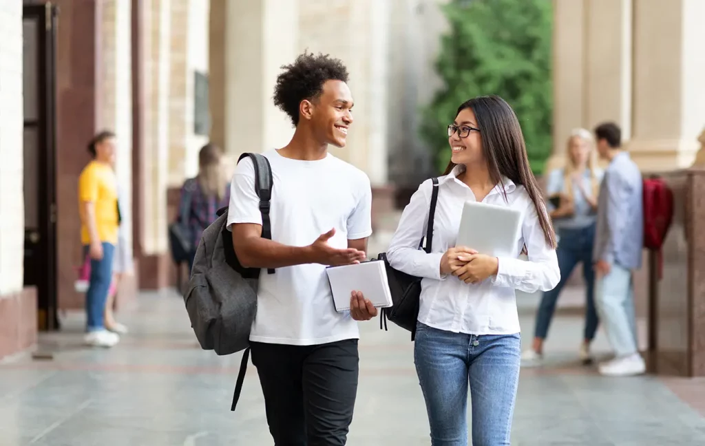 Two friends holding books and wearing back packs talking.