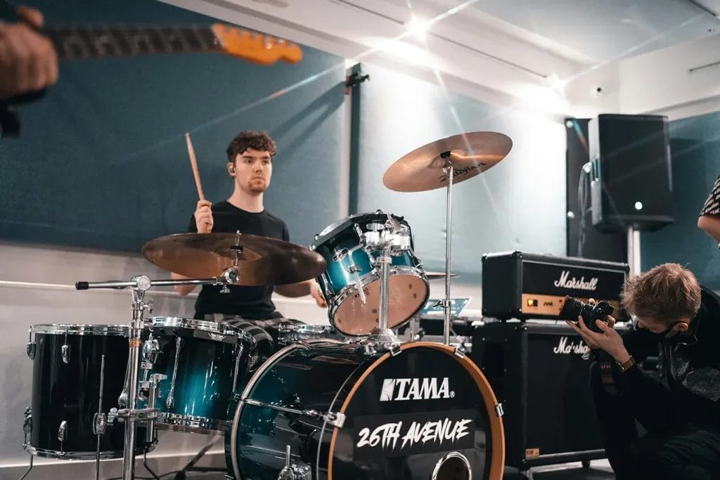 A drummer playing in a recording studio while a photographer takes pictures of them.