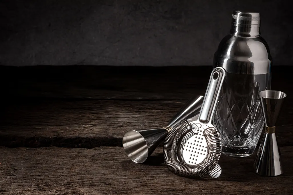 A close-up shot of bartender equipment including a shaker, strainer, and jigger sitting on a wood surface on the right side of the frame.