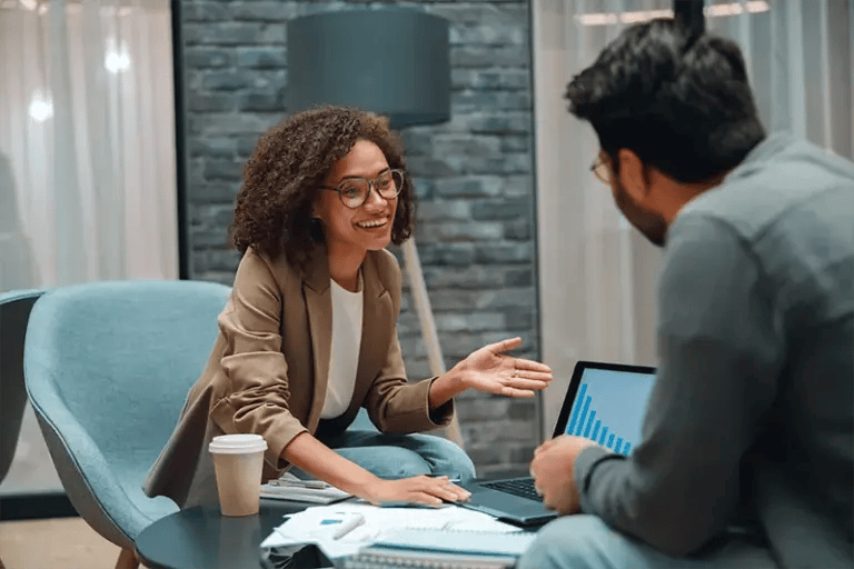A woman with brown curly hair wearing a tan blazer smiles and talks to a man with his back to the camera in a corporate office setting.