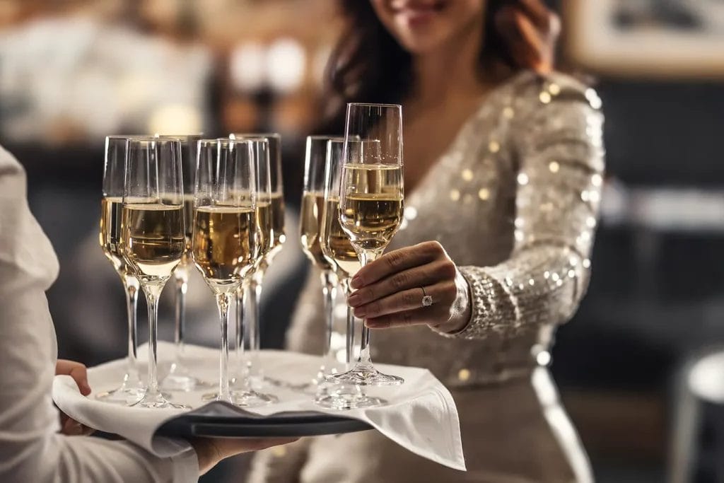A woman in a silver sequined dress takes a champagne flute off of a wedding bartender’s serving tray.