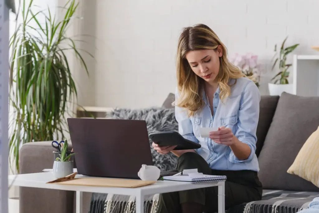 A woman working on her budget at home while sitting on a couch with her laptop on a small table in front of her.
