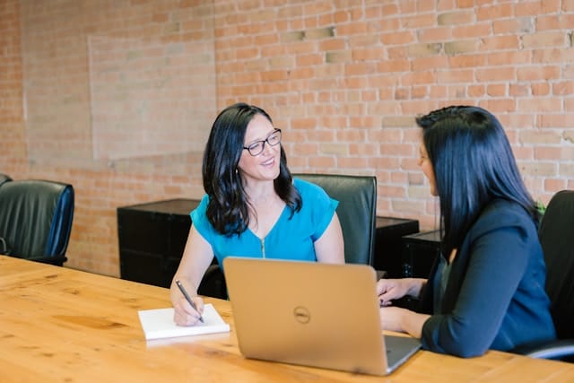 Two women speak at the same table chat next to an open laptop