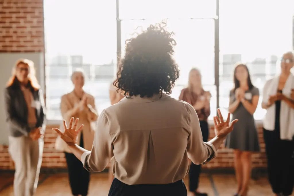 A woman with short, curly hair stands with her back to the camera as she addresses a group of people in a room with large windows during a life coaching event.