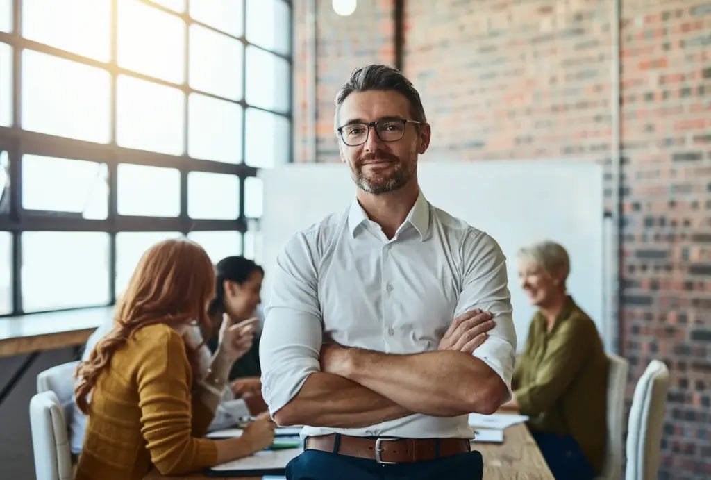Portrait of a confident life coach standing with arms crossed in front of a table of clients. Show less