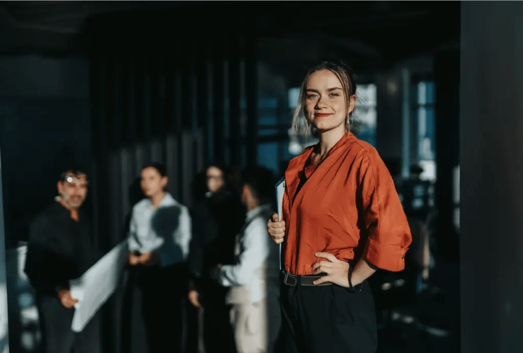 Life coach smiles confidently in the foreground with hand on hip while clients stand in the background in a modern office setting.