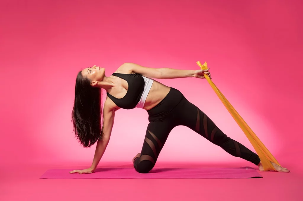 A woman in workout clothes does a yoga pose with a resistance band on a pink yoga mat.