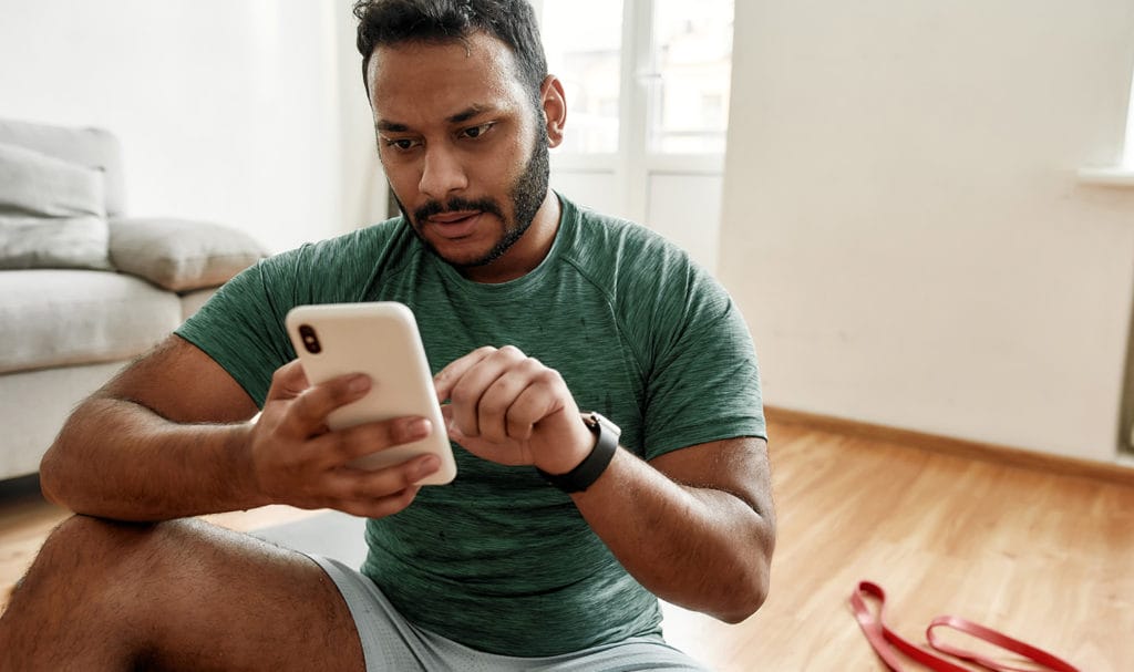 A virtual personal trainer is sitting on the floor of his living room next to his resistance band as he types on his phone.