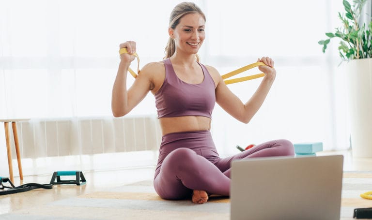 A woman in workout clothes holds a resistance band and smiles while looking at her laptop set up on the floor in front of her.