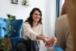 A female life coach in a beige shirt and grey pants smiles and extends her hand to a client sitting in the foreground of the image.