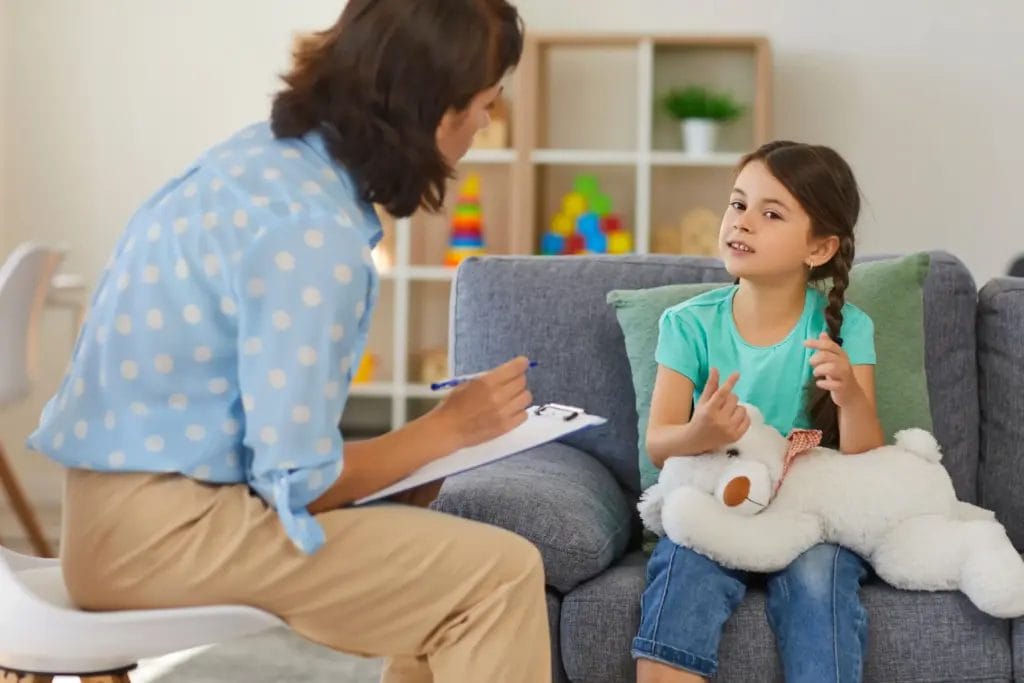 A young girl with braids holds a teddy bear in her lap while talking to a counselor in a blue polka dot shirt.