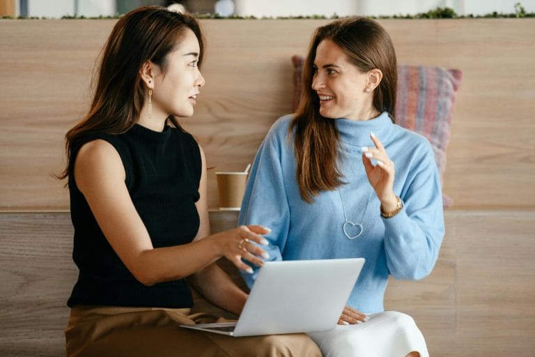 A life coach with her laptop on her lap, sitting next to a client having a discussion at a coffeeshop.