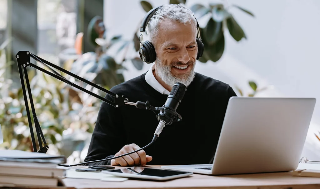 A male business owner smiles as he sits at his computer in his home office with his microphone and headset.