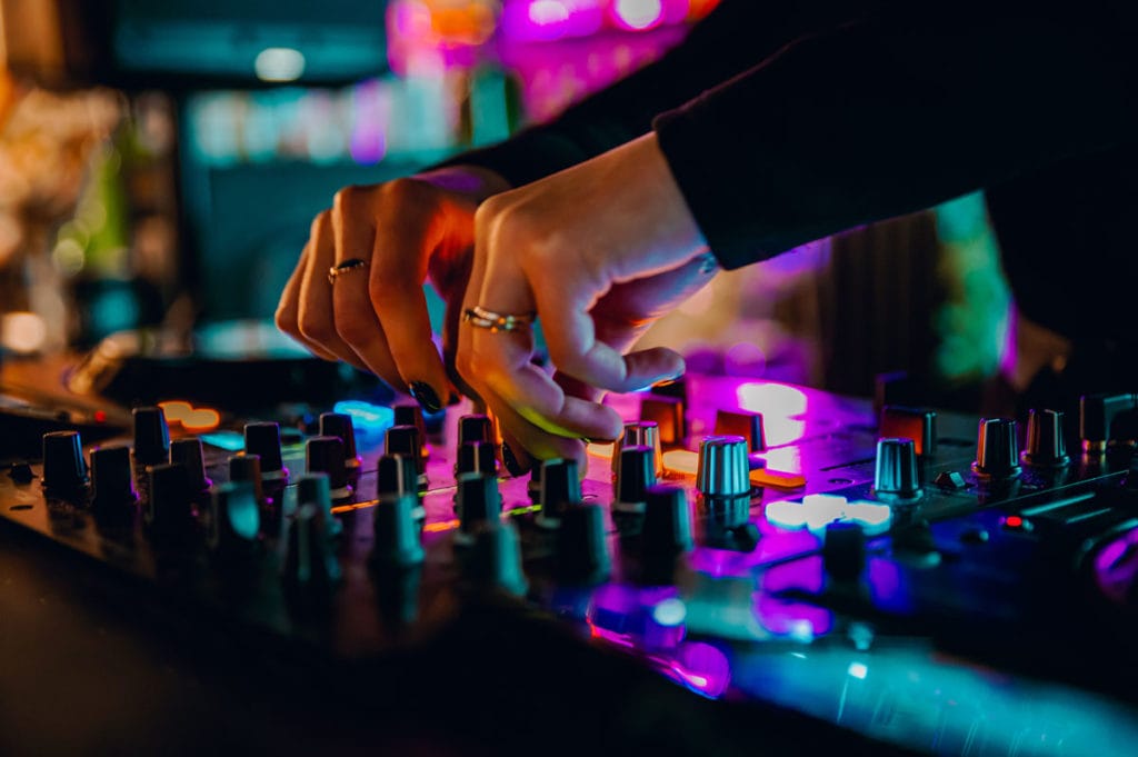 Close-up of a DJ's hands adjusting the controls on a mixing console in a dimly lit setting, with colorful lights reflecting off the equipment.