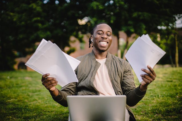 a smiling man sits on a lawn with a computer on his lap, holding papers in both hands