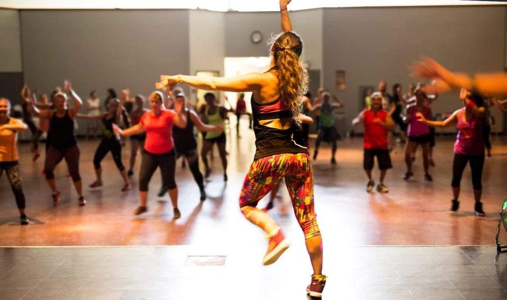 A woman with long wavy hair and colorful clothing, photographed from behind while on a stage, leads a large dance class.