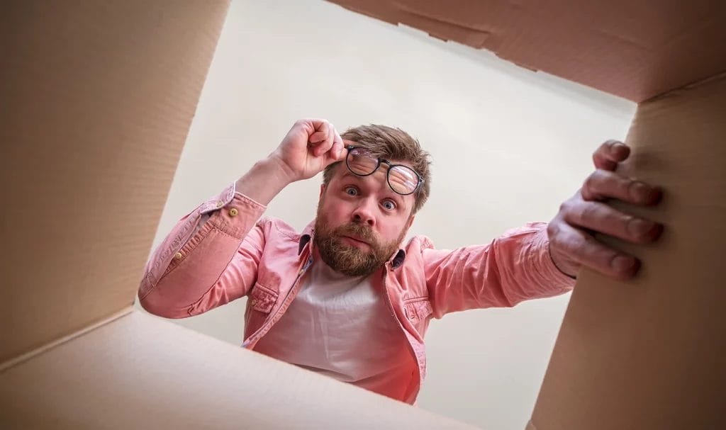 A shot from a box a man is peering down into with a shocked expression on his face as he raises his glasses off his face.