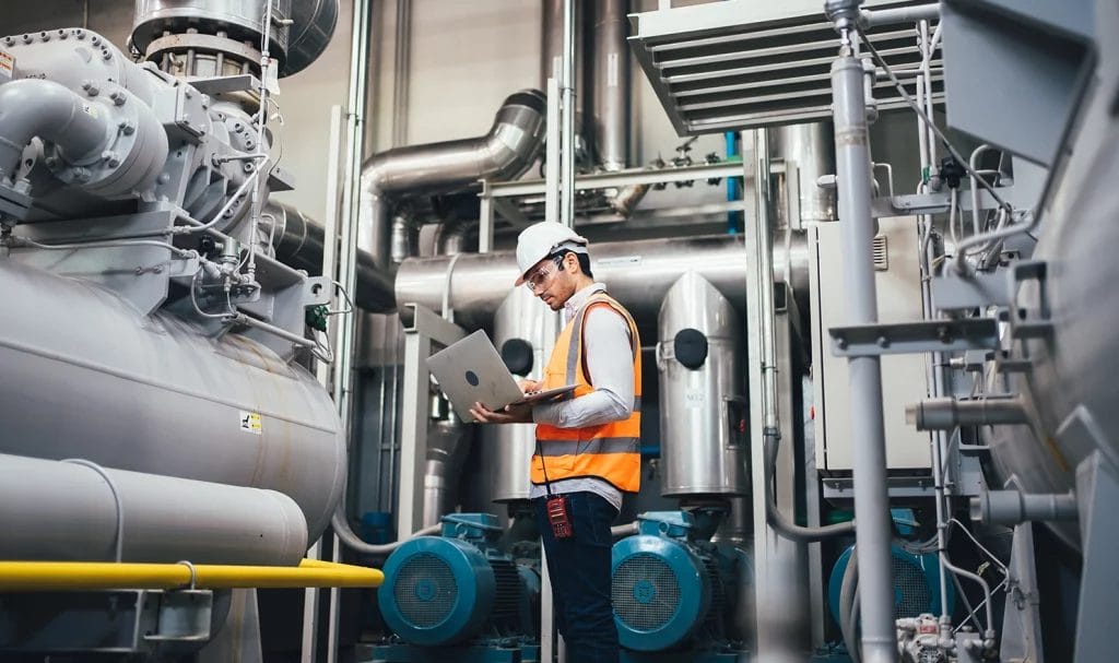 A manufacturering employee wearing a hardhat and high res safety vest is checking on his machinery while carrying a laptop.