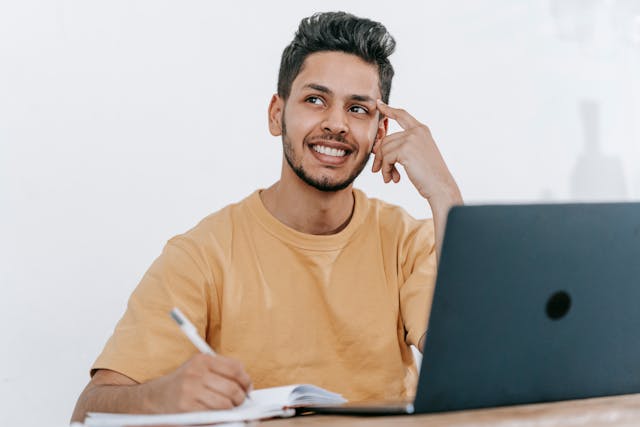 a smiling man sits in front of an open laptop with a pen in hand