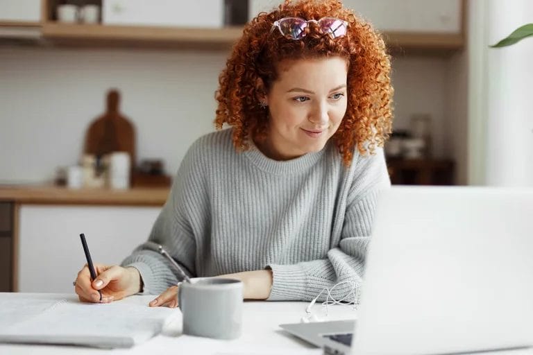 A woman with curly hair, sitting at a desk in front of her laptop taking notes.