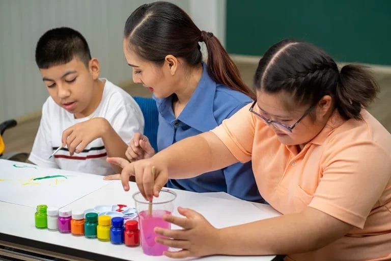 A special education teacher guides two students through a craft project in a classroom.