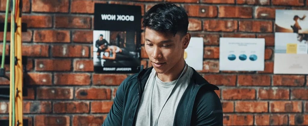 Fitness instructor with black hair, in a gray shirt with a dark gray athletic jacket working on a laptop behind a desk, in front of a brick wall.