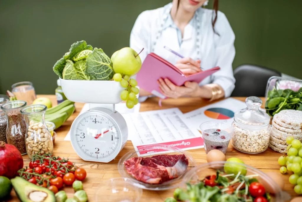 A light brown table featuring a variety of fruits, vegetables, proteins, and other food with a white food scale and a nutritionist wearing white writing in a planner in the background.