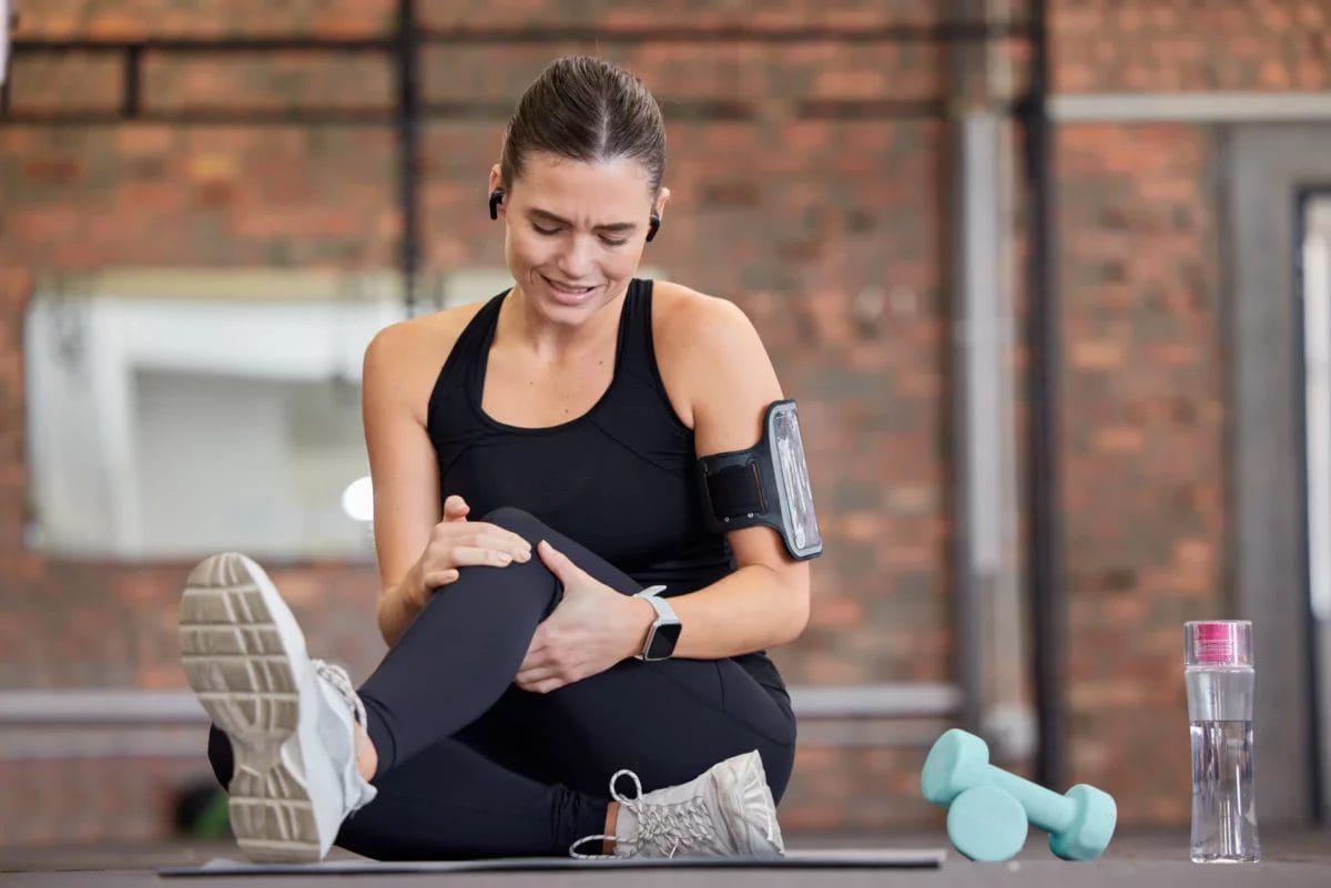 Woman in black workout clothes, sitting on a gym floor holding her left knee in pain.