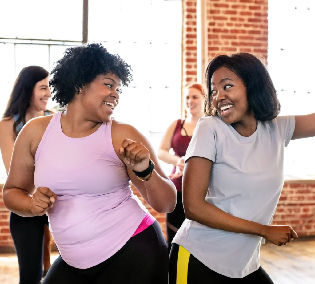 Two women bump hips and smile at each other during a Zumba class in a red brick studio.