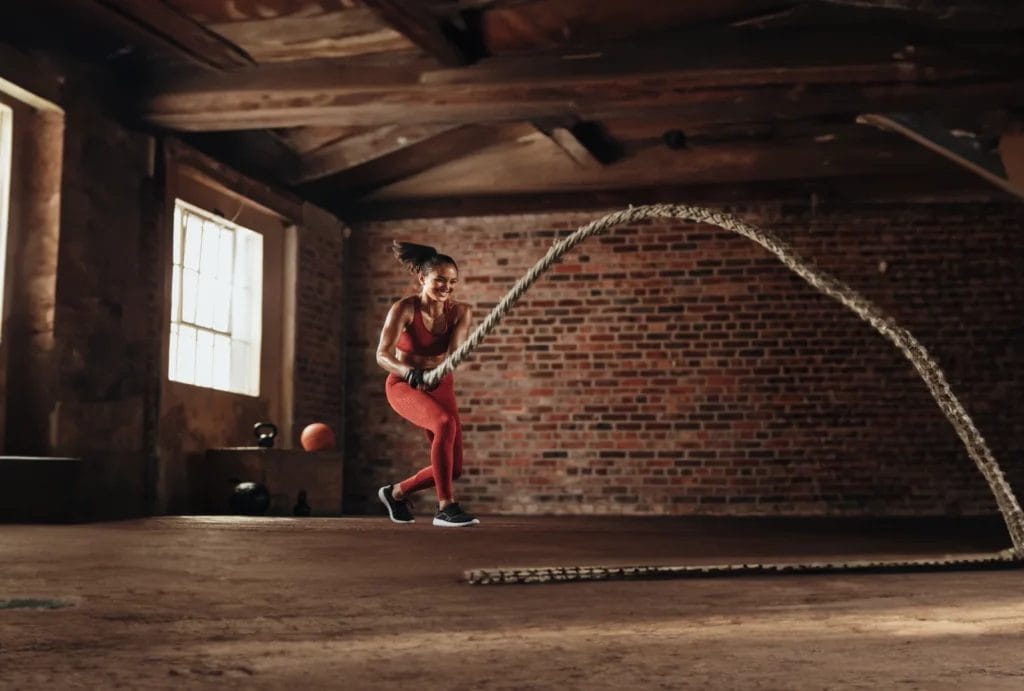 A woman wearing a red workout set smiles and swings a heavy rope during a CrossFit session in a red brick studio.