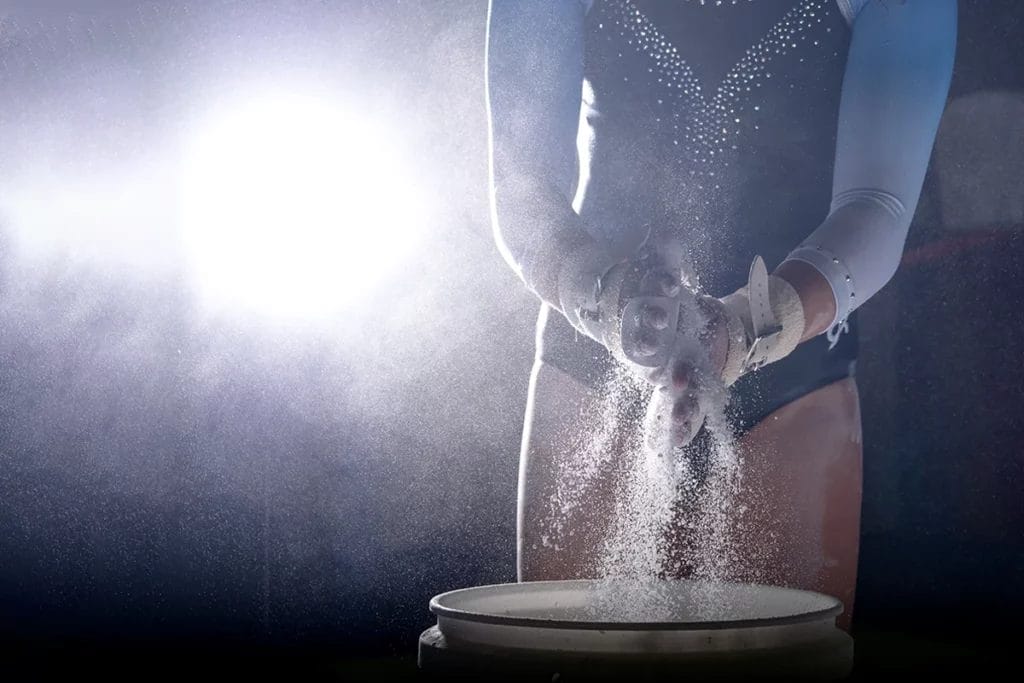 A gymnasts puts chalk on her hands in front of strong back-lighting.