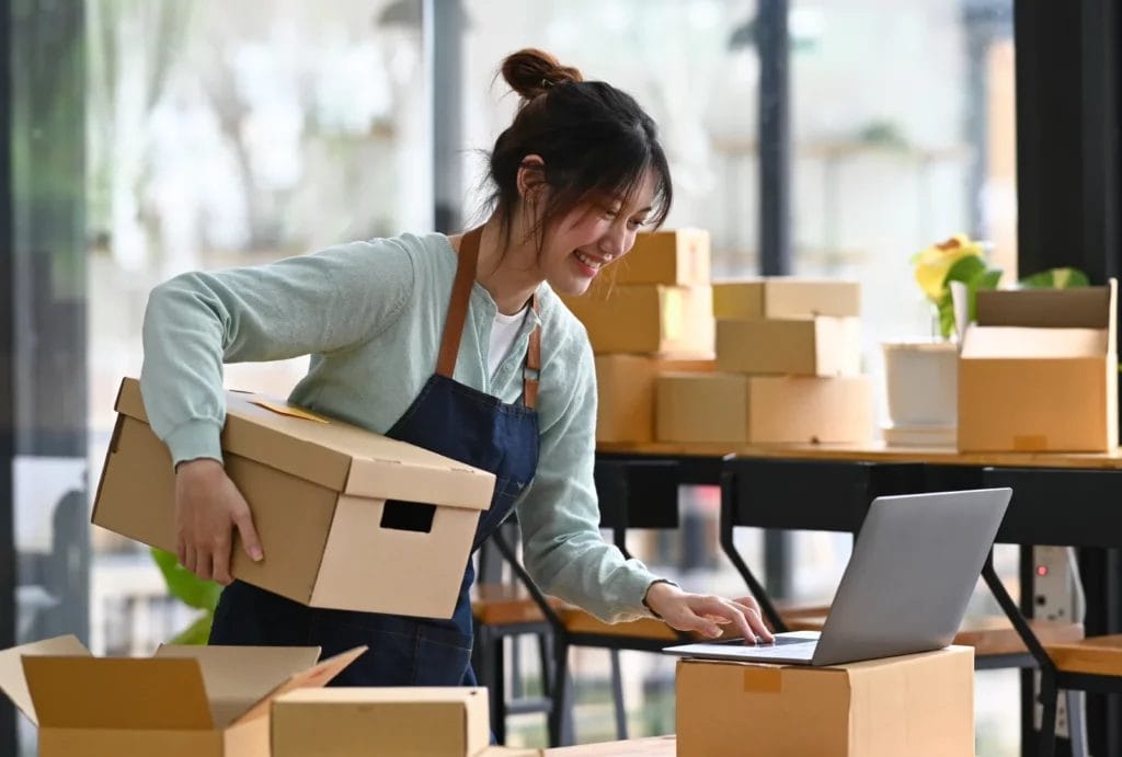 A female small business owner holding a cardboard box and typing on her laptop while packing products for shipment.