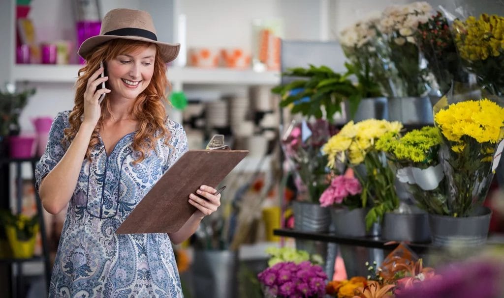 A flower vendor is happily talking on the phone while holding a clipboard next to a stand of flowers.