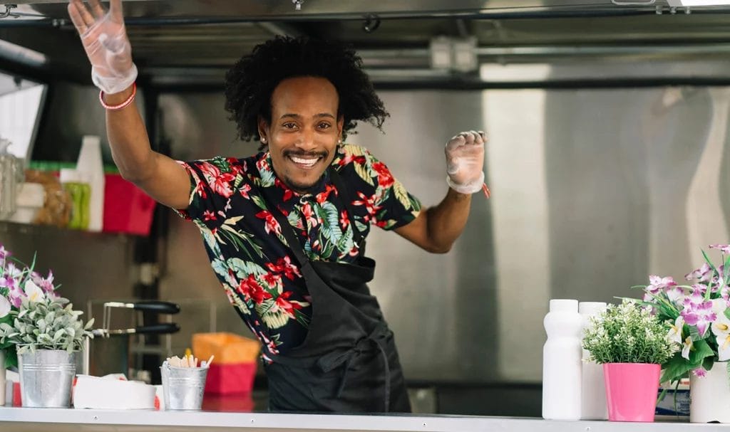 A food truck owner happily smiles and waves from his truck.