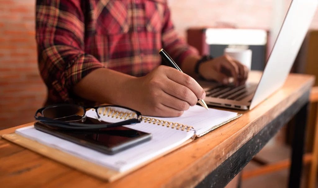 close-up of a small business owner taking notes on a wood desk next to a laptop