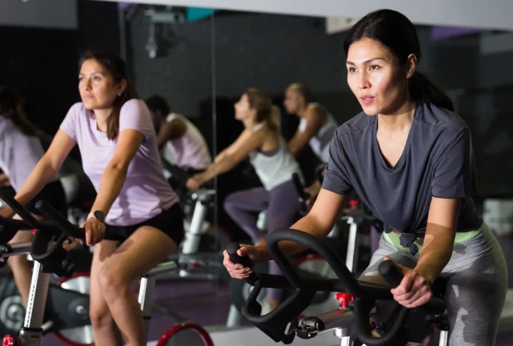Two women in a spin concentrating on their workout, each on a stationary bike, with others in the background adding to the group exercise environment.