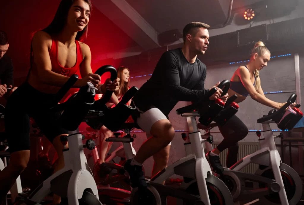 A group of people in a high-energy spin class, riding stationary bikes under dynamic red lighting, creating an intense and engaging atmosphere.