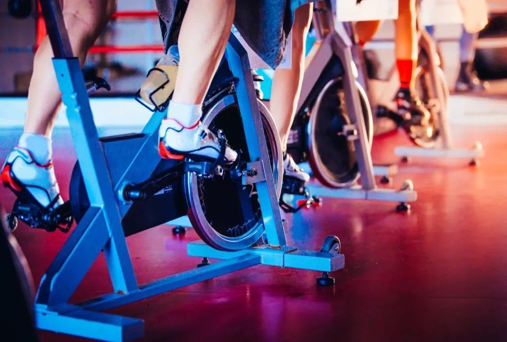 Close-up of individuals’ feet pedaling on spin bikes, showcasing cycling shoes and the intensity of the workout in a spin class setting.