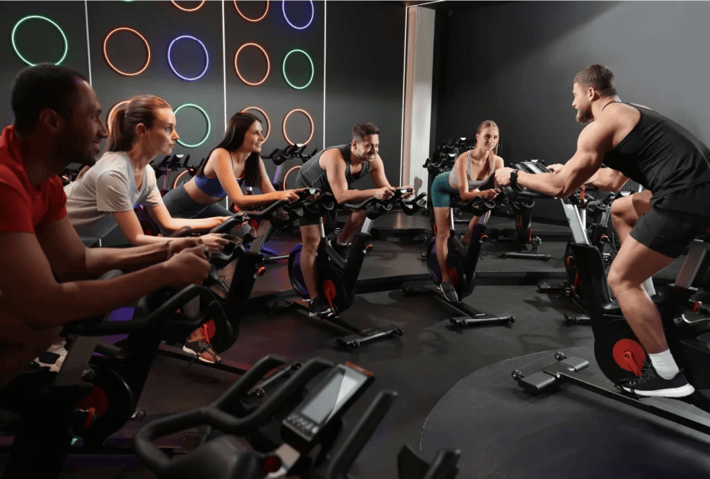 An instructor leading a group spin class in a studio with neon circle lights on the wall, encouraging an interactive and energetic exercise experience.