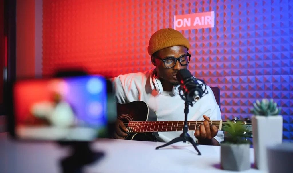 Musician wearing black glasses and a yellow beanie sings into a microphone while playing a guitar in a radio station, backlit by red and blue light.