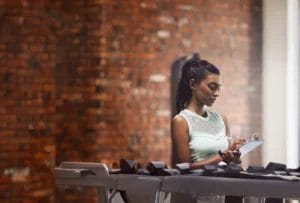 A young woman with long black hair in a ponytail wearing a mesh mint-colored sleeveless top takes notes on her clipboard while assessing the weight rack in a gym.